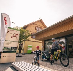 Das Mobilitätszentrum in Münsingen im Biosphärengebiet Schwäbische Alb. Zwei Erwachsene und ein Kind stehe in kurzer Radkleidung mit ihren Fahrrädern vor einem Gebäude. Die beiden Erwachsenen haben E-Bikes und einer einen Kinderfahrradanhänger. Vor dem Gebäude steht ein Schild mit der Aufschrift Mobilitätszentrum und eine Beach Flag von Bosch.