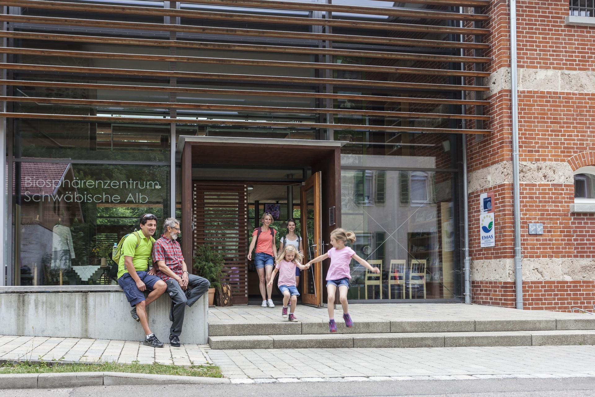 Das Biosphärenzentrum Schwäbische Alb in Münsingen im Biosphärengebiet. Zwei Personen sitzen auf einer Steinmauer vor dem Biosphärenzentrum Schwäbische Alb. Aus der Tür kommen  lächelnd zwei Erwachsene, zwei Kinder springen die Stufen vor dem Eingang hinunter.