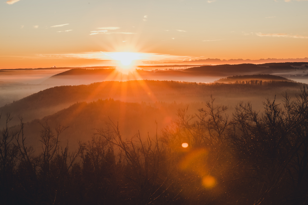 Aussicht vom Sternbergturm in der Nähe von Münsingen im Biosphärengebiet Schwäbische Alb. Eine Hügelllandschaft, von der lediglich die Silhoutte erkennbar ist. Dazwischen sind Nebelschwaden. Die Sonne wirft lange Strahlen und taucht das gesamte Bild in ein strahlendes orangefarbenes Licht.