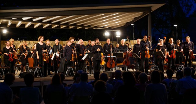 Das Sommernachtskonzert in Münsingen im Biosphärengebiet Schwäbische Alb. Ein Orchester aus einigen Musikern*innen mit verschiedenen Instrumenten steht auf einer beleuchteten Bühne. Davor sitzen im Dunkeln mehrere Personen.