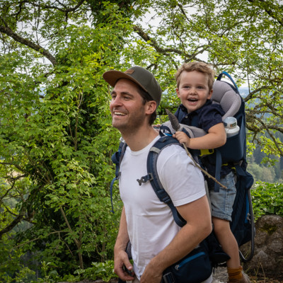 Familienwanderung mit Burgen im Großen Lautertal in Münsingen im Biosphärengebiet Schwäbische Alb. Ein Erwachsener hat eine Kraxe mit einem Kind auf dem Rücken. Beide lachen herzhaft. Im Hintergrund ist eine Mauer und ein großer Baum.