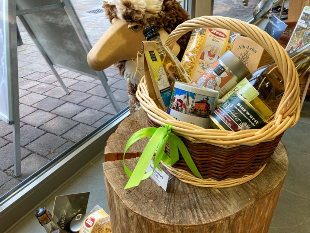 Ein Geschenkkorb in der der Touristik Information Münsingen. Auf einem Holzstamm steht ein geflochtener Korb mit mehreren regionalen Produkten darin. Daneben ist ein Deko-Schaf.