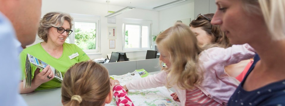 Die Touristik Information Münsingen im Biosphärengebiet Schwäbische Alb berät seine Gäste gerne zu allen möglichen Themen. Eine Person steht hinter einem Thresen und zeigt mehreren Erwachsenen und Kindern Landkarten.