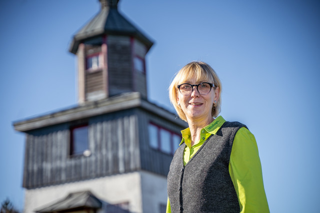 Diana Krause von der Touristik Information Münsingen auf dem ehemaligen Truppenübungsplatz  im Biosphärengebiet Schwäbische Alb. Eine Person mit Brille, blonden Haaren, grüner Bluse und grauer Weste lächelt in die Kamera. Im Hintergrund ist verschwommen ein Turm.