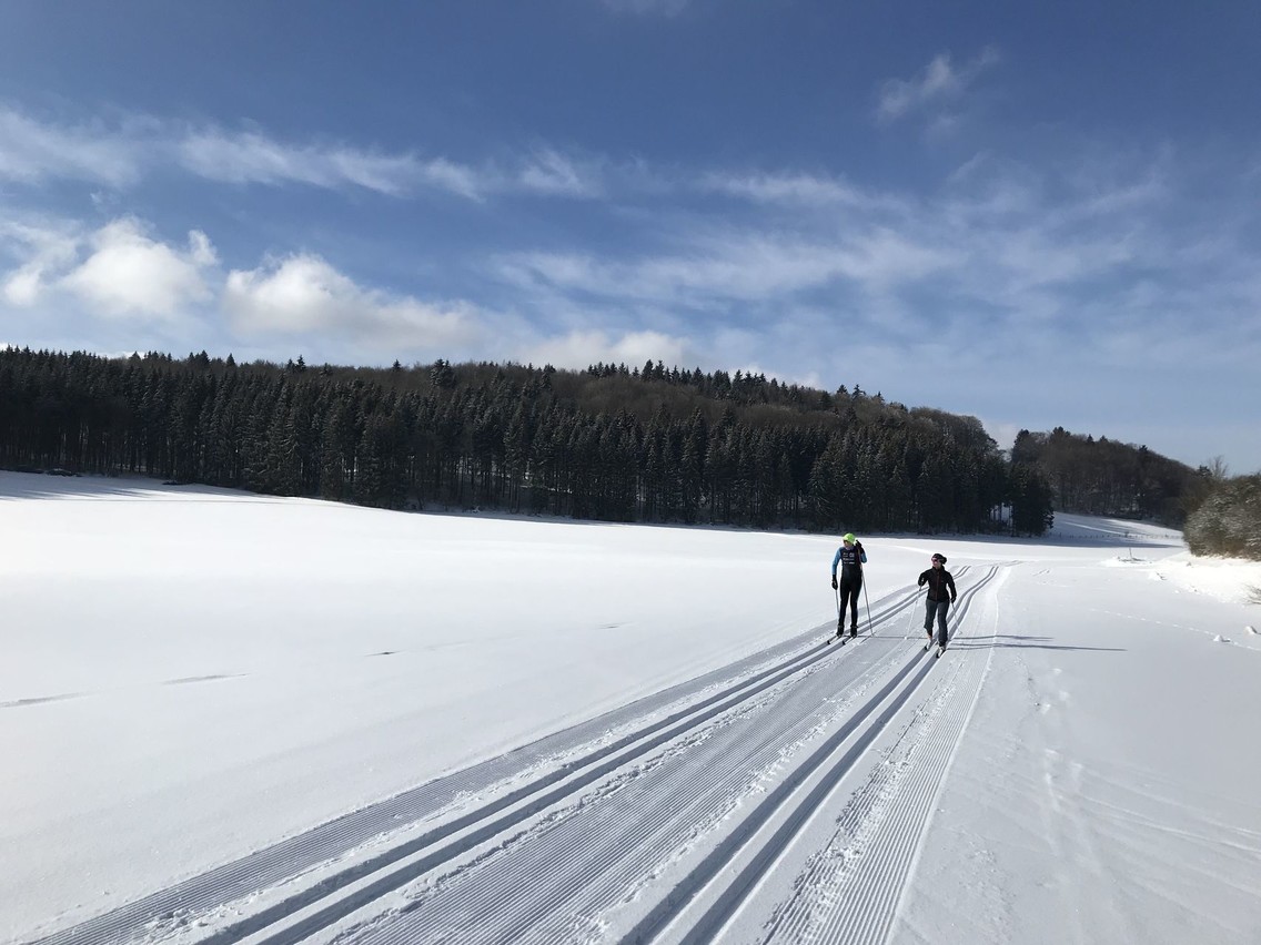 Klassische Langlaufloipe in Münsingen im Biosphärengebiet Schwäbische Alb. Zwei klassische Langläufer laufen nebeneinander in den Langlaufspuren. Die Landschaft ist mit Schnee bedeckt und im Hintergrund ist Wald.