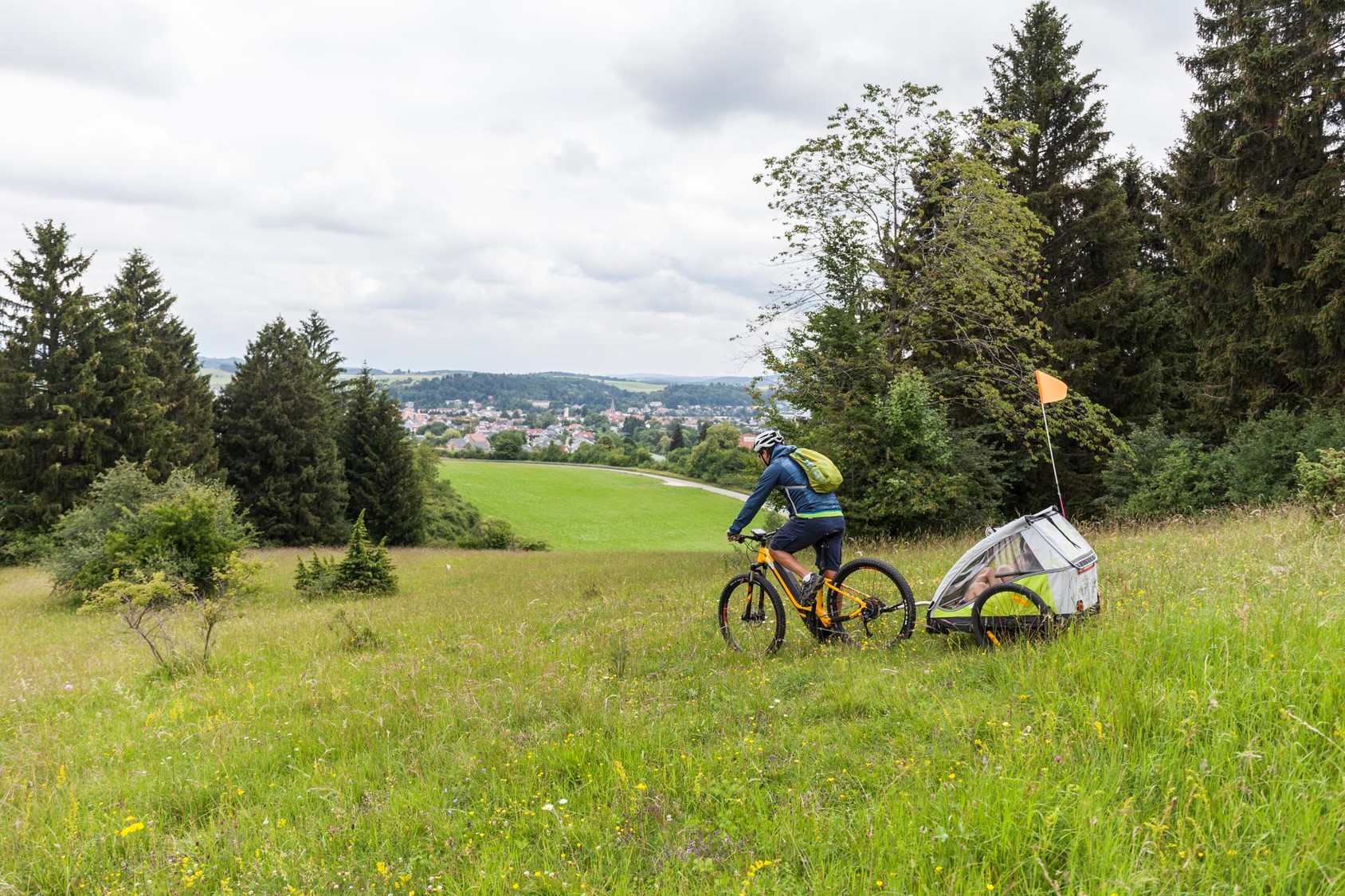 Familienradtour in Münsingen im Biosphärengebiet Schwäbische Alb. Ein*e Radfahrer*in fährt durch eine Sommerwiese. Hinten angehängt ist ein Kinderanhänger mit Fahne. Links und rechts ist Wald. Davor ist ein Ausblick über Wiesen und eine Stadt.