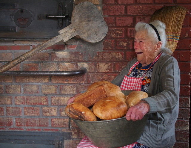 Backhaus in Münsingen im Biosphärengebiet Schwäbische Alb. Eine alte Frau mit weißen Haaren lächelt fröhlich und hält eine Blechwanne, gefüllt mit Brot, in ihrem Schoß. Hinter ihr ist eine Backsteinwand mit einer Klappe.