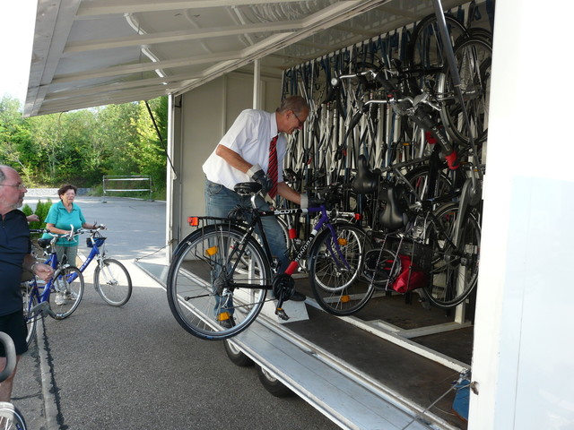 Busverkehr mit Fahrradtransport ins Große Lautertal in Münsingen im Biosphärengebiet Schwäbische Alb. Eine Person mit Hemd und Krawatte lädt ein Fahrrad in einen Anhänger, in dem weitere Fahrräder hängen. Drumherum stehen Radfahrer*innen mit ihren Bikes.
