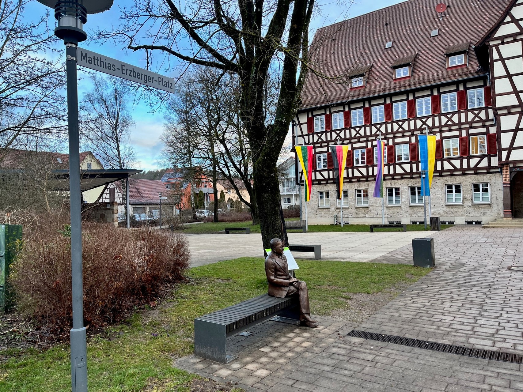 Der Matthias-Erzberger-Platz in Münsingen im Biosphärengebiet Schwäbische Alb. Auf einer Bank sitzt eine bronzefarbene Statue. Davor ist ein Platz mit einem großen historischen Fachwerkgebäude, vor dem verschiedene Fahnen wehen. Links ist eine Laterne mit dem Schild und der Aufschrift Matthias-Erzberger-Platz.