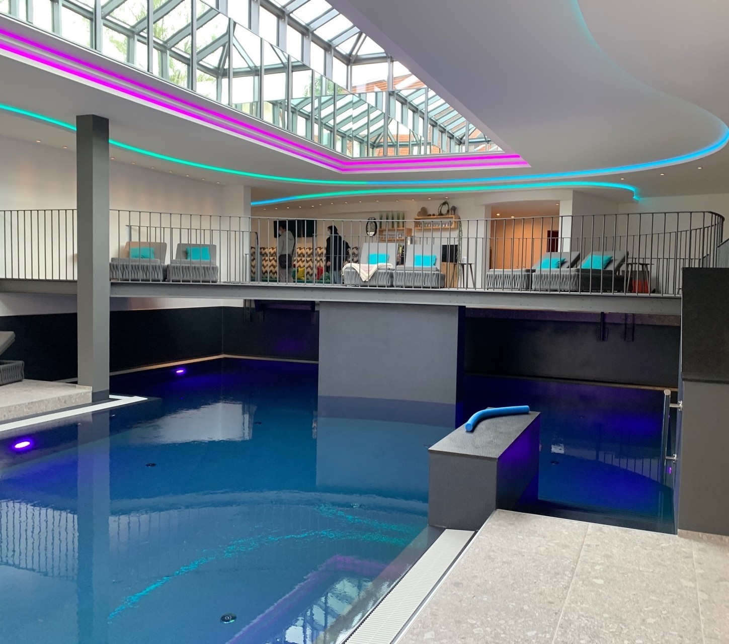 Wellness im Biosphärenhotel Gasthof Herrmann in Münsingen im Biosphärengebiet Schwäbische Alb. Ein blauer Pool, dessen Wasser ruhig steht und der stimmungsvoll beleuchtet ist. An der Decke sind wietere stimmungsvolle Lichter in blau und pink. Über dem Pool ist eine Terrasse mit Liegen.