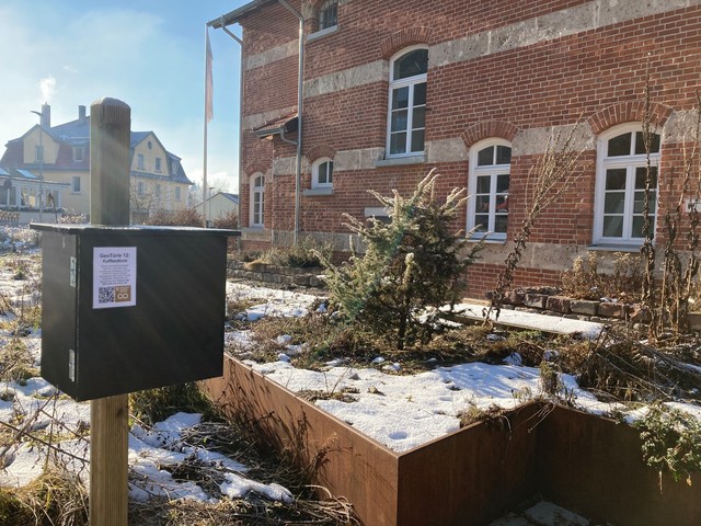 GeoTürle am Biosphärenzentrum in Münsingen im Biosphärengebiet Schwäbische Alb. An einem Holzpfosten hängt ein Briefkasten mit einer längeren Beschriftung. Dahinter ist ein Beet, das mit Schnee bedeckt ist. Im Hintergrund ist ein Backsteinhaus. Die Sonne scheint.