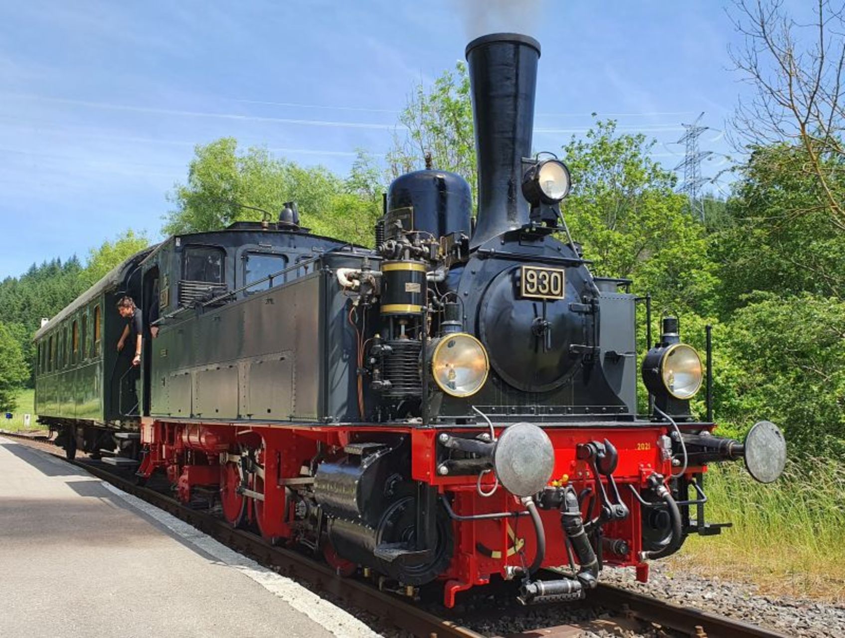 Nostalgiefahrten mit der Dampflok der Schwäbische Alb-Bahn in Münsingen im Biosphärengebiet Schwäbische Alb. Eine historische schwarze Dampflok mit einem grünen Waggon steht auf Gleisen an einem Bahnhof. Aus der Fahrerkabine lehnt eine Person. Es ist sonnig.