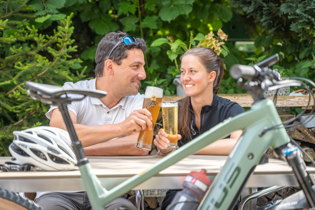 Die Berg Bier-Tour in Münsingen im Biosphärengebiet Schwäbische Alb. Zwei Personen sitzen in einem Biergarten und stoßen mit Berg-Bier an. Davor steht ein e-Bike.