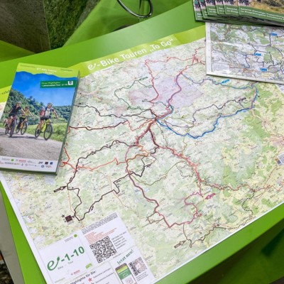 Münsingen auf der CMT auf der Messe Stuttgart. Auf einem grünen Tisch liegt eine Karte mit mehreren eingezeichneten Wegen in verschiedenen Farben. Daneben liegt ein Flyer mit Radfahrer*innen darauf und ein weiterer Flyer mit einer Karte.