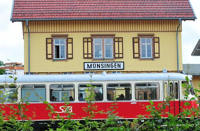Der Bahnhof mit der Schwäbischen Alb-Bahn in Münsingen im Biosphärengebiet Schwäbische Alb. Im Vordergrund ein roter Schienenbus, der teilweise vo einigen Ästen mti grünen Blättern verdeckt wird, dahinter ein historisches gelbes Gebäude