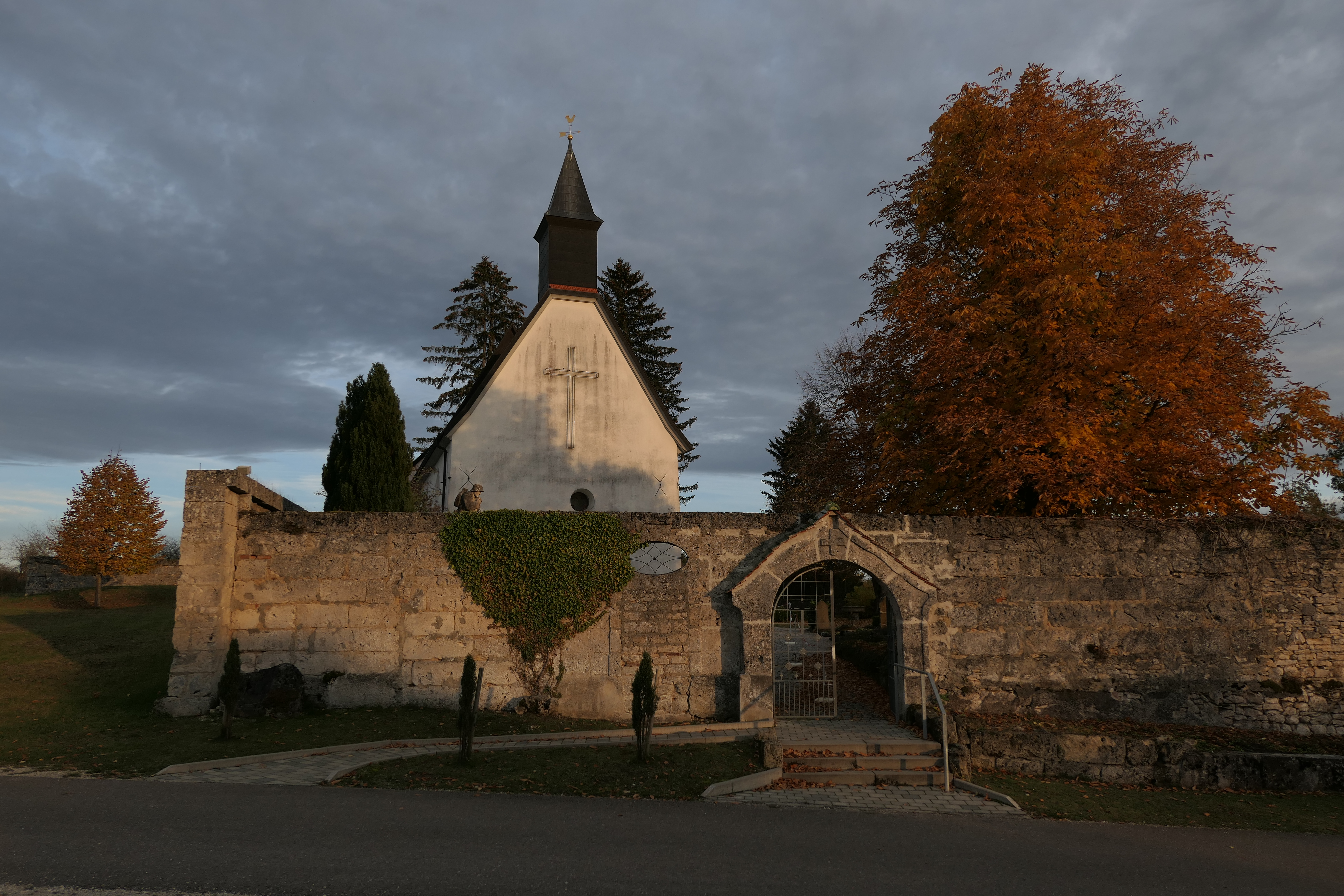 Gruorn im Herbst auf dem ehemaligen Truppenübungsplatz Münsingen im Biosphärengebiet Schwäbische Alb. Eine Kirche mit einem großen Kreuz auf der Fassade. Davor eine Steinwand mit einem tor. Ringsherum einige Bäume mit bunten Blättern. Der Himmel ist bedeckt.
