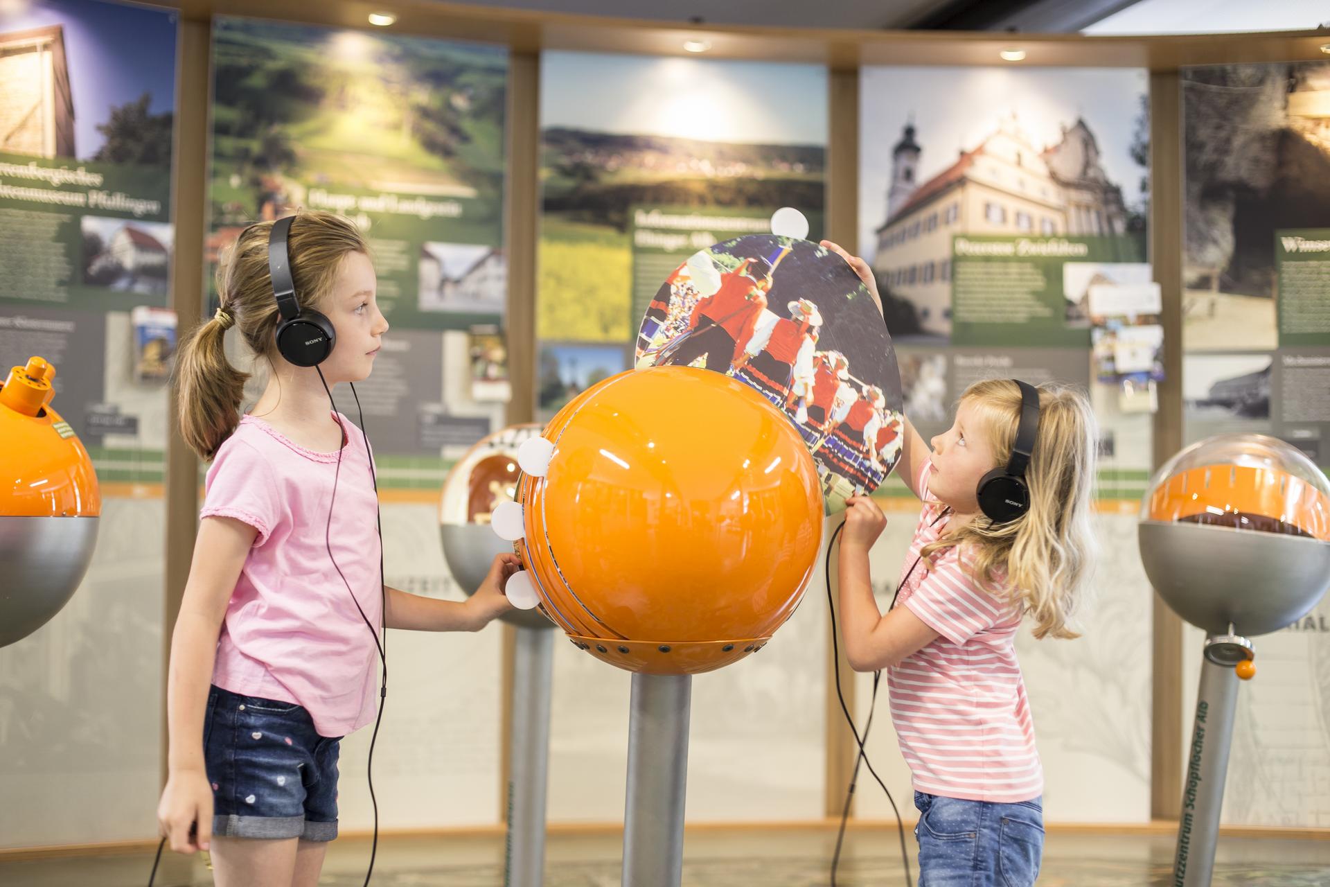 Ein Familienausflug ins Biosphärenzentrum Schwäbische Alb in Münsingen im Biosphärengebiet Schwäbische Alb. Zwei Kinder stehen mit Kopfhörern an einem interaktiven Ausstellungsstück: Eine orangefarbene Kugel, aus der ein Kind ein Bild herauszieht. Im HIntergrund sind weitere Kugeln und Erklärtafeln.