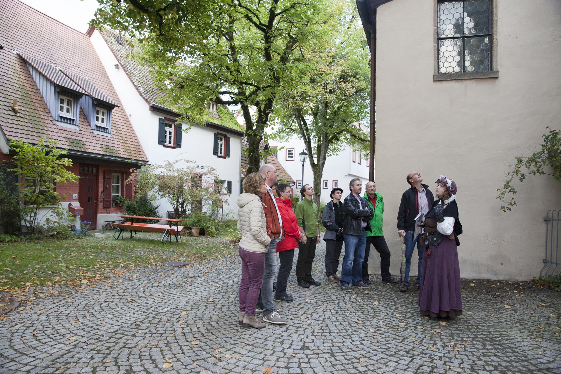 Stadtführung mit dem Marktweib Marga in Münsingen im Biosphärengebiet Schwäbische Alb. Eine Person in einem mittelalterlichen Kostüm steht vor einer Gruppe und erzählt. Die Gruppe hört ihr zu. Sie stehen auf einem kleinen Platz. Hinter ihnen sind kleine Häuser und zwei Bäume.