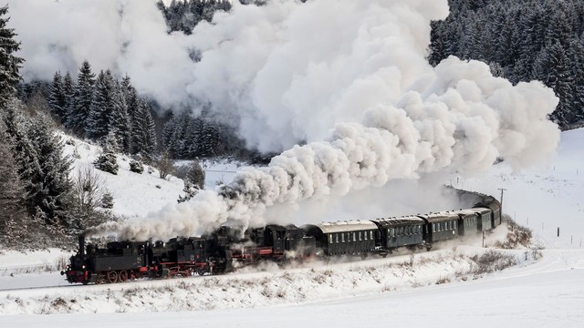 Nostalgiefahrten mit dem Dampfzug in Münsingen im Biosphärengebiet Schwäbische Alb. Eine Eisenbahn fährt durch die verschneite Winterlandschaft. Aus dem Kamin kommt Qualm, der weit hinter dem Zug hinterher zieht. Im Hintergrund ist ein Tannenwald.